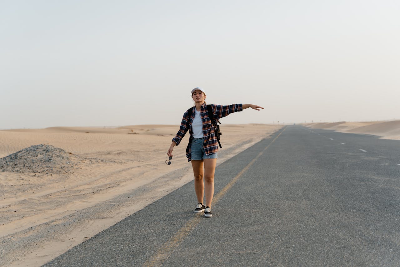 A young woman with a backpack hitchhiking on an open desert road under clear skies.