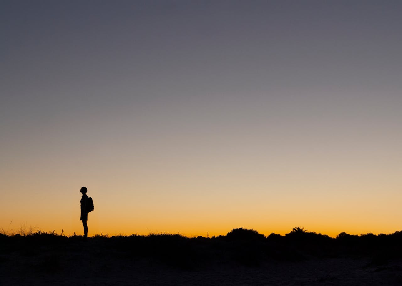 A lone figure stands silhouetted against a vibrant orange sunset sky, creating a tranquil outdoor scene.